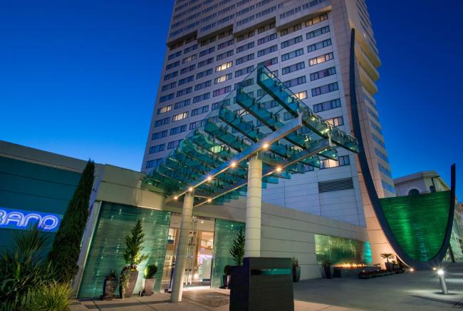 The image shows a modern building with a glass canopy at its entrance, lit up in the evening. The architecture features clean lines and green accents.